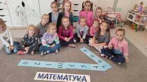 Dziewczynki siedzą w półkolu na szarym dywanie. Przed nimi są ułożone cyfry: 1 i napis matematyka.