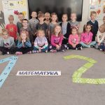 Dzieci siedzą w półkolu na szarym dywanie. Przed nimi są ułożone cyfry: 1 i 2 oraz napis matematyka.