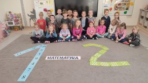 Dzieci siedzą w półkolu na szarym dywanie. Przed nimi są ułożone cyfry: 1 i 2 oraz napis matematyka.