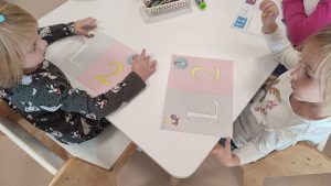 Dzieci siedzą przy białych stolikach i kolorują kredkami cyfrę 2 i literę L.