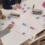 Dzieci siedzą przy białych stolikach i kolorują kredkami cyfrę 2 i literę L.