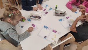 Dzieci siedzą przy białych stolikach i kolorują kredkami cyfrę 2 i literę L.