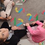 Przed grupką dzieci, które siedzą na szarym dywanie leżą ułożone z klocków NEMI cyfry 1 i 2.