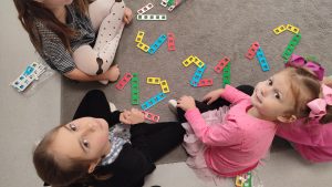 Przed grupką dzieci, które siedzą na szarym dywanie leżą ułożone z klocków NEMI cyfry 1 i 2.