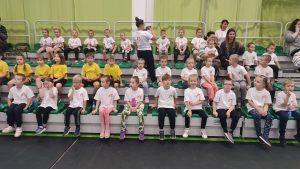 Grupka dzieci siedzi na zielonych krzesełkach na trybunie sali gimnastycznej.
