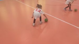 Dziewczynka turla zieloną piłkę po podłodze w hali sportowej.