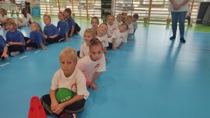 10 dzieci w białych koszulkach siedzą w rzędzie i czekają na swoją kolej w zawodach sportowych.