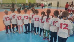 10 dzieci w białych koszulkach z cyfrą 12 stoją plecami i oglądają wręczanie nagród.