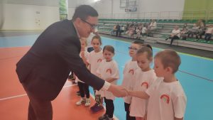 Mężczyzna wręcza grupie dzieci dyplom uczestnictwa w zawodach sportowych i gratuluje poszczególnym dzieciom.