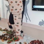 Na zdjęciu chłopiec z pomocą Pani przechodzący boso przez ścieżkę sensoryczną z liści, kasztanów, szyszek, żołędzi.