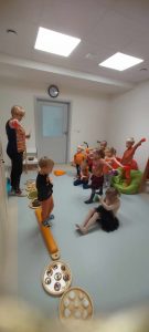 Dzieci stojąc przed nauczycielem, wykonują ćwiczenia gimnastyczne.