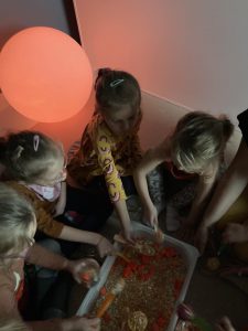 Zabawa sensoryczna. Dzieci siedzą w kółeczku. Za pomocą łyżeczek z plastikowego pojemnija, wkładają do słloiczków różnej wielkości pomarańczowe pomponiki.