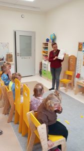 Dzieci siedzą na krzesełkach. Słuchają mamy jednego z dzieci, która przedstawia swój zawód.