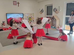 Grupa dzieci tańcząca na środku sali z materiałami w kolorach białych i czerwonych. W tle, pod ściana siedzą dzieci. Z prawej strony stoi pani nauczycielka.