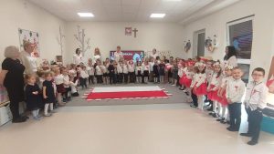 Dzieci oraz panie odświętnie ubrane, śpiewające hymn. Na środku, na sali, na dywanie leży flaga biało-czerwona