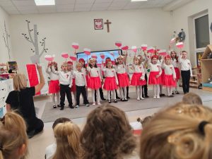 Grupa dzieci ubranych na galowo stojąca w dwóch rzędach i trzymająca w rękach mapę Polski w barwach biało-czerwonych.