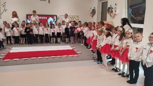 Grupy dzieci ubrane na galowo, śpiewające hymn Polski. Za dziećmi stoją cztery panie. Przed dziećmi, na dywanie leży flaga biało-czerwona. Wokół flagi leżą mapy Polski w barwach biało-czerwonych.