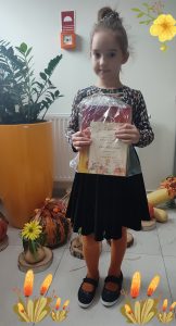 Dziewczynka w czarnej spódniczce i bluzce w brązowe ciapki , trzyma w ręce nagrodę za udział w konkursie wokalnym. Dziewczynka pozuje do zdjęcia.