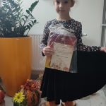Dziewczynka w czarnej spódniczce i bluzce w brązowe ciapki , trzyma w ręce nagrodę za udział w konkursie wokalnym. Dziewczynka pozuje do zdjęcia.