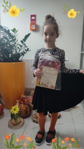 Dziewczynka w czarnej spódniczce i bluzce w brązowe ciapki , trzyma w ręce nagrodę za udział w konkursie wokalnym. Dziewczynka pozuje do zdjęcia.