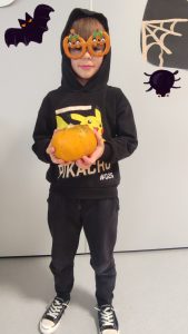 Chłopiec ubrany na czarno w okularach i kapturze na głowie trzyma w ręce dynię i pozuje do zdjęcia z symbolami Halloween.