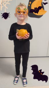 Dziewczynka ubrana na czarno trzyma w ręce dynię i pozuje do zdjęcia z symbolami Halloween.