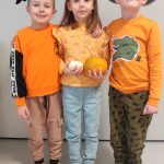 Jedna dziewczynka i dwóch chłopców ubranych na pomarańczowo pozują do zdjęć z symbolami Halloween.