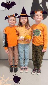 Jedna dziewczynka i dwóch chłopców ubranych na pomarańczowo pozują do zdjęć z symbolami Halloween.