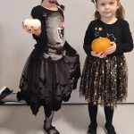Dwie dziewczynki ubrane w czarne stroje czarownic trzymają w ręce dynie. Pozują do zdjęcia, które ozdobione jest symbolami Halloween.