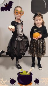 Dwie dziewczynki ubrane w czarne stroje czarownic trzymają w ręce dynie. Pozują do zdjęcia, które ozdobione jest symbolami Halloween.