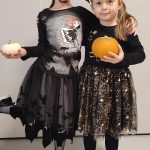 Dwie dziewczynki ubrane w czarne stroje czarownic trzymają w ręce dynie. Pozują do zdjęcia, które ozdobione jest symbolami Halloween.