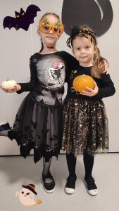 Dwie dziewczynki ubrane w czarne stroje czarownic trzymają w ręce dynie. Pozują do zdjęcia, które ozdobione jest symbolami Halloween.
