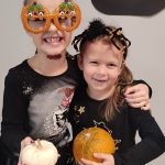 Dwie dziewczynki ubrane w czarne stroje czarownic trzymają w ręce dynie. Pozują do zdjęcia, które ozdobione jest symbolami Halloween.