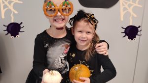 Dwie dziewczynki ubrane w czarne stroje czarownic trzymają w ręce dynie. Pozują do zdjęcia, które ozdobione jest symbolami Halloween.