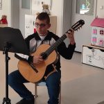 Gitarzysta Tobiasz gra na gitarze. Przed nim leżą nuty. Naokoło siedzą dzieci i słuchają muzyki.
