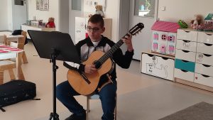 Gitarzysta Tobiasz gra na gitarze. Przed nim leżą nuty. Naokoło siedzą dzieci i słuchają muzyki.