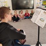 Gitarzysta Tobiasz gra na gitarze. Przed nim leżą nuty. Naokoło siedzą dzieci i słuchają muzyki.