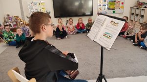 Gitarzysta Tobiasz gra na gitarze. Przed nim leżą nuty. Naokoło siedzą dzieci i słuchają muzyki.