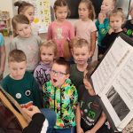 Gitarzysta Tobiasz siedzi na krześle i trzyma na kolanach gitarę. Pokazuje dzieciom jak się gra na gitarze. Obok stoją dzieci.