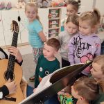 Gitarzysta Tobiasz siedzi na krześle i trzyma na kolanach gitarę. Pokazuje dzieciom jak się gra na gitarze. Obok stoją dzieci.