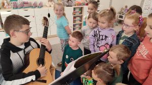 Gitarzysta Tobiasz siedzi na krześle i trzyma na kolanach gitarę. Pokazuje dzieciom jak się gra na gitarze. Obok stoją dzieci.