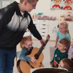 Dziewczynka siedzi na krześle i trzyma na kolanach gitarę. Obok stoją dzieci.