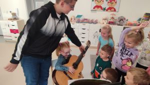 Dziewczynka siedzi na krześle i trzyma na kolanach gitarę. Obok stoją dzieci.