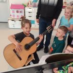Chłopiec siedzi na krześle i trzyma na kolanach gitarę. Obok stoją dzieci.