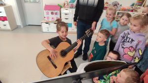 Chłopiec siedzi na krześle i trzyma na kolanach gitarę. Obok stoją dzieci.