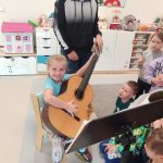 Dziewczynka siedzi na krześle i trzyma na kolanach gitarę. Obok stoją dzieci.