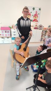 Dziewczynka siedzi na krześle i trzyma na kolanach gitarę. Obok stoją dzieci.