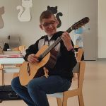 Gitarzysta Tobiasz gra na gitarze. Przed nim leżą nuty. Naokoło siedzą dzieci i słuchają muzyki.