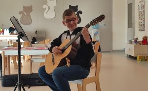 Gitarzysta Tobiasz gra na gitarze. Przed nim leżą nuty. Naokoło siedzą dzieci i słuchają muzyki.