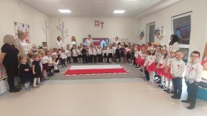 Cztery grupy dzieci stoją naokoło flagi biało-czerwonej i śpiewają hymn narodowy.